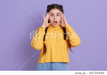 Stunned dark haired woman with two pigtails, keeping hands near face, afraids of something astonishing, dresses in fashionable yellow shirt, posing isolated over lilac background. 89794680