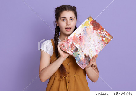 Astonished woman with painting palette in hands, keeps mouth opened, looks surprised, standing in casual clothing against lilac wall, expressing astonish. Astonished woman with painting palette in hands, keeps mouth opened, looks surprised, standing in casual clothing against lilac wall, expressing astonish. 89794746