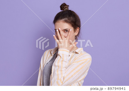 Attractive lady covering her face with fingers, woman with dark hair and bun, female posing isolated over lilac background, girl wearing white casual shirt. Attractive lady covering her face with fingers, woman with dark hair and bun, female posing isolated over lilac background, girl wearing white casual shirt. 89794941