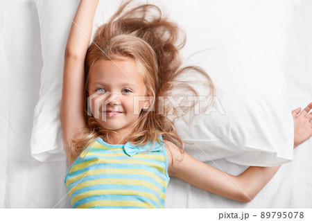 Above view of attractive light haired small child, has blue eyes, stretches in bed, lies on white pillow, dressed in colourful striped pyjamas, enjoys recreation time in bedroom. Children and rest 89795078