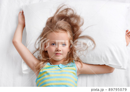 View from above of beautiful small lovely female child with light hair, dressed in striped pyjamass, lies on pillow, has good rest in bed, awakes early in morning. Childhood and leisure concept. View from above of beautiful small lovely female child with light hair, dressed in striped pyjamass, lies on pillow, has good rest in bed, awakes early in morning. Childhood and leisure concept. 89795079