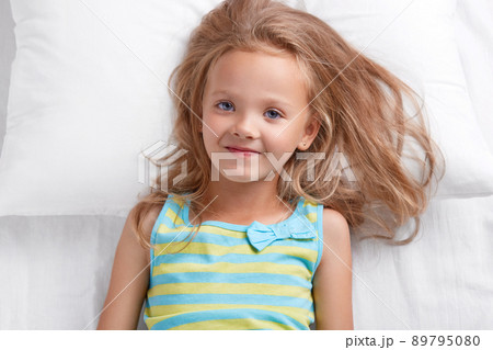Photo of pleasant looking small female kid with blue eyes, dressed in nightwear, has satisfied expression, lies on white bedclothes, enjoys good rest, rejoices starting new day. Bed time and children 89795080