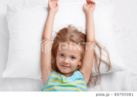 Top view of pretty child has long hair, stretches in bed, lies on white pillow, has good mood after rest in bed, awakes early in morning, wants to start new day, looks directly at camera. Childhood 89795081