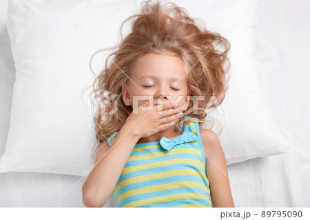 Pretty sleepy beautiful small female girl keeps eyes closed, palm on mouth, dressed in casual striped nightwear, lies on comfortable bed, awakes after sleeping. Children, good morning concept Pretty sleepy beautiful small female girl keeps eyes closed, palm on mouth, dressed in casual striped nightwear, lies on comfortable bed, awakes after sleeping. Children, good morning concept 89795090