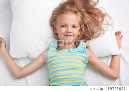 Children, bedding and awakening concept. Pleased small European girl with charming tender smile, long light hair, dressed in pyjamass, lies on white pillow and bedclothes, has appealing appearance. Children, bedding and awakening concept. Pleased small European girl with charming tender smile, long light hair, dressed in pyjamass, lies on white pillow and bedclothes, has appealing appearance. 89795091