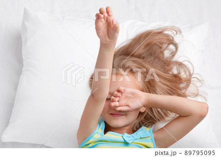 Photo of small adorable cute child covers eyes, stretches in bed, dressed in pyjamass, lies on comfortable bed, doesnt want to get up, enjoys good rest and dreams. Childhood and sleeping concept 89795095