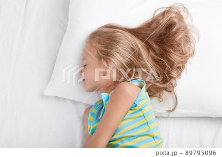 Sideways shot of small kid wears nightwear, being deep in sleep, rests at bed, lies on white bed, enjoys bedtime, rests after active games outdoor with friends, naps in bedroom. Children concept 89795096