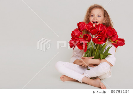 Excited little girl of school age sitting on floor with crossed legs and embracing bouquet of red tulips, having happy expression, copy space for promotion, isolated over gray background. 89795134