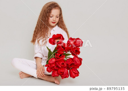 Gentle blonde girl with wavy hair sitting on floor and looking at red tulips in her hands, having dreamy expression, copy space for advertisement, isolated over gray background. Gentle blonde girl with wavy hair sitting on floor and looking at red tulips in her hands, having dreamy expression, copy space for advertisement, isolated over gray background. 89795135