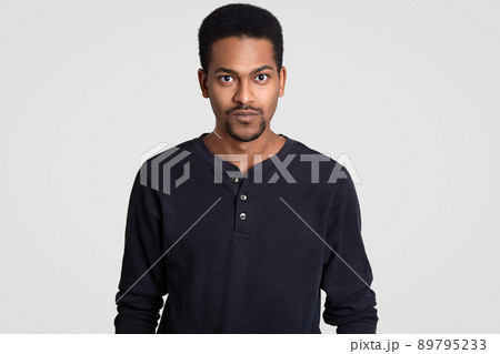 Indoor shot of serious self confident dark skinned young man with curly short hair, looks with stern expression, models indoor against white background. People, ethnicity and lifestyle concept 89795233