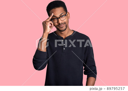 Dark skinned handsome male looks thoughtful. Man wears glasses and black sweater. Young guy with beard keeps fore finger on forehead. Model poses in photo studio isolated over rose background. 89795257