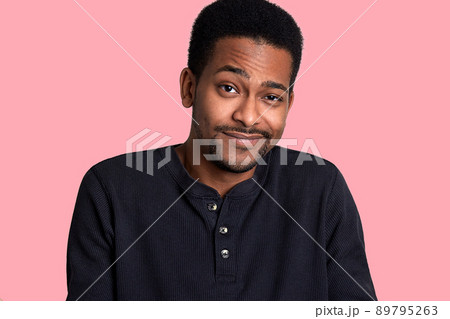 African man with beard and mustache crews his eyes and smiles, male looks shy. Model stands isolated over pink blackground, poses and looks at camera. Young guy wears black shirt. People concept. 89795263