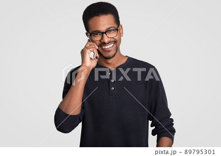 Positive dark skinned Afro American man with cheerful expression, has telephone conversation, discusses something pleasant with friend, organize party together, isolated over white background. 89795301