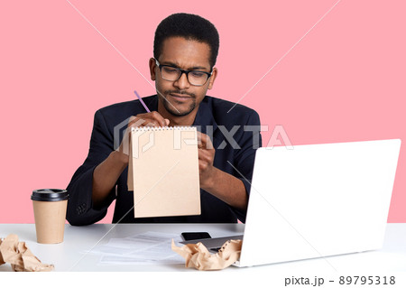 Thoughtful black businessman tries to write poem in notebook for her girlfriend while working on laptop. Handsom African American guy wears blak shirt and jacket sits against pink wall in studio. 89795318