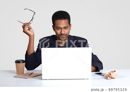 Photo of serious dark skinned man works freelance, keeps eyewear in hands, reads news on internet website, focused into monitor of laptop, wears formal suit, sits in coworking space over white wall 89795329