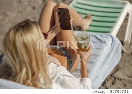 Young woman lying on deckchair with smartphone and cocktail outdoors 89796731