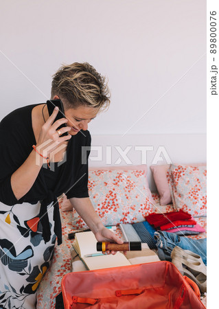 mature woman traveller using a smart phone and packing her suitcase preparing to travel on a summer holiday. Vertical mature woman traveller using a smart phone and packing her suitcase preparing to travel on a summer holiday. Vertical 89800076