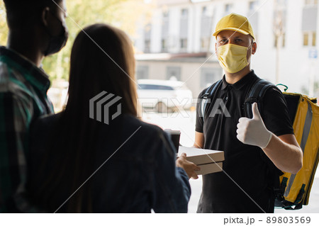 Smilling delivery man in yellow face mask, uniform cap delivering cardboard boxes with pizza 89803569