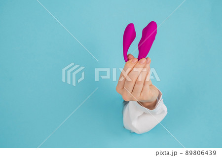 A woman's hand holds a pink sex toy through a hole in a paper blue background. A woman's hand holds a pink sex toy through a hole in a paper blue background. 89806439