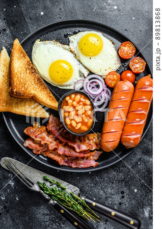 English breakfast with fried eggs, sausages, bacon, beans and toasts in a plate. Black background. top view 89813868
