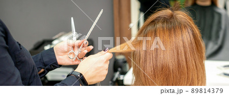 Red-haired woman sitting a front of the mirror and receiving haircut her red long hair by a female hairdresser in a hair salon, back view Red-haired woman sitting a front of the mirror and receiving haircut her red long hair by a female hairdresser in a hair salon, back view 89814379