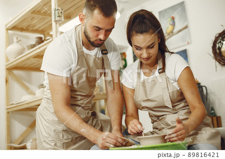 Mutual creative work. Young beautiful couple in casual clothes and aprons. People creating a bowl on a pottery wheel in a clay studio. Mutual creative work. Young beautiful couple in casual clothes and aprons. People creating a bowl on a pottery wheel in a clay studio. 89816421