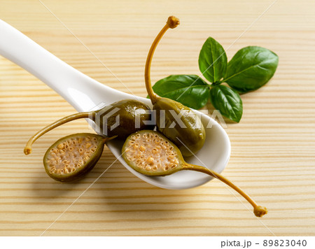Pickled caper berries in a white spoon and basil leaf on a wooden background. Pickled caper berries in a white spoon and basil leaf on a wooden background. 89823040