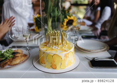Family celebrating happy birthday on summer terrace. Beautiful yellow birthday cake. Background of people eating dinner. Food concept outdoors. Different kind of food and drinks on the table 89824128