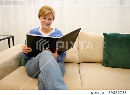 Happy charming middle-aged Caucasian woman sits on a comfortable sofa in the lobby of a beauty salon and leafs through a magazine while waiting for an appointment with a aesthetician Happy charming middle-aged Caucasian woman sits on a comfortable sofa in the lobby of a beauty salon and leafs through a magazine while waiting for an appointment with a aesthetician 89826783