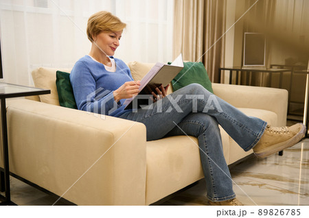 Charming middle-aged European woman sits on a comfortable couch in the lounge area of a wellness spa clinic and leafs through a catalog while waiting for an appointment with a doctor cosmetologist 89826785