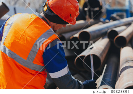 Builder in helmet and vest measures diameter of pipe. Workflow. Open warehouse with metal pipes. Builder in helmet and vest measures diameter of pipe. Workflow. Open warehouse with metal pipes. 89828392