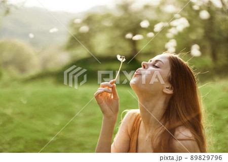 The happy woman smiles and blows the dandelion in the wind. Summer green landscape and sunshine in the background The happy woman smiles and blows the dandelion in the wind. Summer green landscape and sunshine in the background 89829796