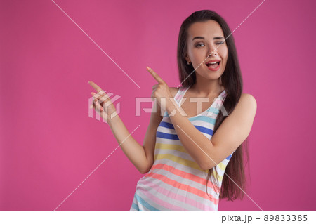 Brunette woman with long hair, dressed in colorful striped shirt, posing against pink studio background. Sincere emotions. Close-up. 89833385