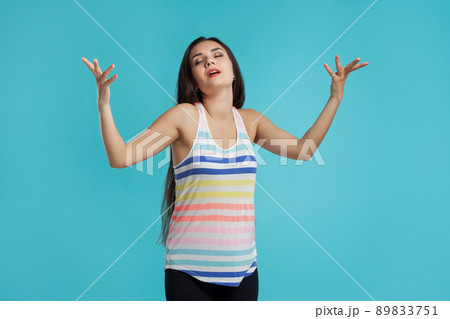 Brunette woman with long hair, dressed in striped shirt and black leggings, posing against blue studio background. Sincere emotions. Close-up. 89833751