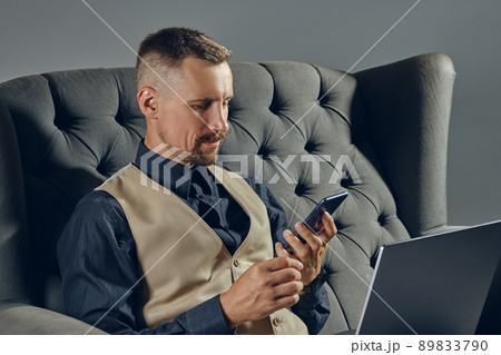 Man with stylish mustache, dressed in black shirt and beige vest is sitting on dark sofa, using his laptop and smartphone. Grey background, close-up. 89833790