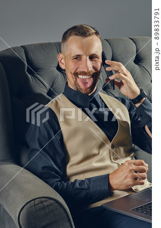 Man with stylish mustache, dressed in black shirt and beige vest is sitting on dark sofa, using his laptop and smartphone. Grey background, close-up. Man with stylish mustache, dressed in black shirt and beige vest is sitting on dark sofa, using his laptop and smartphone. Grey background, close-up. 89833791