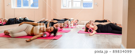 A group of sportswomen doing pilates or yoga on pink mats in a beige loft studio interior. Teamwork, good mood and healthy lifestyle concept. A group of sportswomen doing pilates or yoga on pink mats in a beige loft studio interior. Teamwork, good mood and healthy lifestyle concept. 89834099