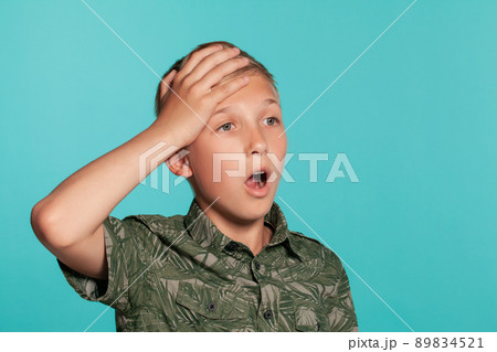 Close-up portrait of a blonde teenage boy in a green shirt with palm print posing against a blue studio background. Concept of sincere emotions. Close-up portrait of a blonde teenage boy in a green shirt with palm print posing against a blue studio background. Concept of sincere emotions. 89834521