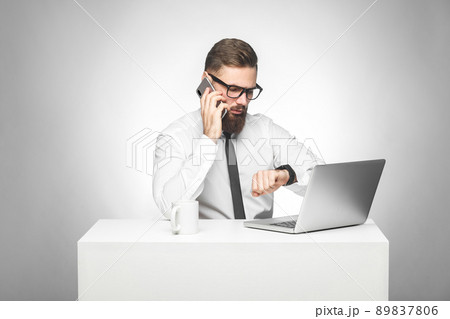 Thoughful bearded young worker in white shirt and black tie are sitting in office on desk and talking with partner on phone also checking time on his own hand watch, planning meeting. Gray background 89837806
