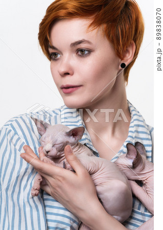 Beautiful redhead young woman hugging Sphynx Cat and looking away. Studio shot on white background. Portrait of hipster with short hair dressed in striped white-blue casual shirt. Part of series. 89838070