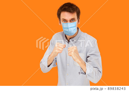 Portrait of angry young worker man with surgical medical mask standing in boxing fists and looking at camera and ready to attack against virus. indoor studio shot isolated on orange background. 89838243