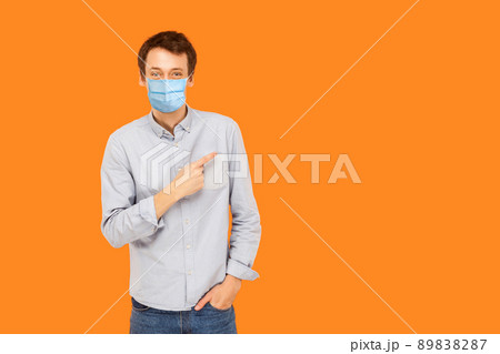 Portrait of happy young worker man with surgical medical mask standing, pointing and showing background empty copy space and smiling. indoor studio shot isolated on orange background. 89838287