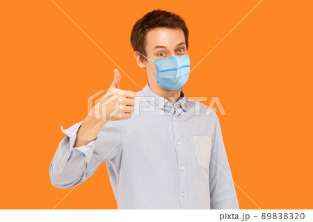 Like. Portrait of satisfied young worker man with surgical medical mask standing thumbs up and looking at camera smiling. indoor studio shot isolated on orange background. Like. Portrait of satisfied young worker man with surgical medical mask standing thumbs up and looking at camera smiling. indoor studio shot isolated on orange background. 89838320