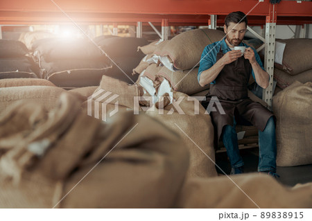 Man barista inhaling aroma of coffee sitting on warehouse near bags with roasted coffee beans 89838915