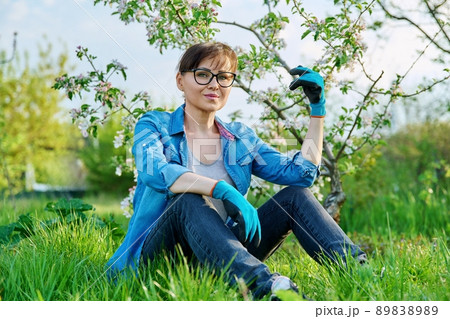 Positive beautiful mature female gardener sitting on the grass with blossoming apple tree. Positive beautiful mature female gardener sitting on the grass with blossoming apple tree. 89838989