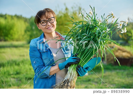 Woman in gardening gloves with white daffodil bush plant and shovel, female looking at camera Woman in gardening gloves with white daffodil bush plant and shovel, female looking at camera 89838990