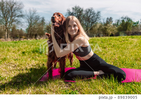 Cute young caucasian woman is meditating while sitting on a rug on the lawn on a sunny summer with Irish Setter dog. 89839005
