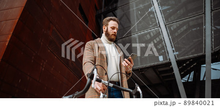 Young man with bicycle holding a smartphone front of building. Creative businessman in a modern business area. Young man with bicycle holding a smartphone front of building. Creative businessman in a modern business area. 89840021