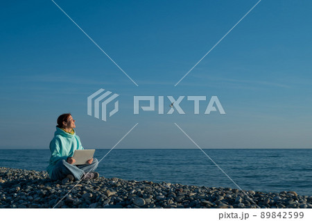 Happy caucasian woman working on a laptop while sitting on a pebble beach. 89842599
