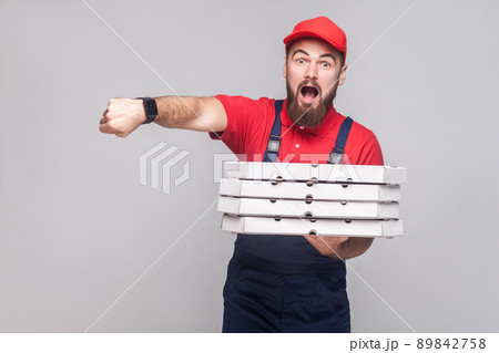 Hurry up! time is gone. Young amazed delivery man with beard in blue uniform and red t-shirt, holding stack of cardboard pizza boxes on grey background. Indoor, studio shot, isolated, copy space. Hurry up! time is gone. Young amazed delivery man with beard in blue uniform and red t-shirt, holding stack of cardboard pizza boxes on grey background. Indoor, studio shot, isolated, copy space. 89842758